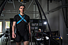 Mann mit schwarzer Sportkleidung und blauem Brustgurt geht in einem Labor mit technischer Ausrüstung und Monitoren im Hintergrund.