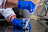 Hände mit blauen Handschuhen halten einen Becher mit Flüssigkeit und einem darin eingetauchten Schlauch in einem Laborumfeld.