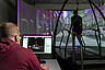Person sitzt vor Computer mit 3D-Analyse auf Bildschirm und beobachtet eine weitere Person, die auf einem Laufband vor einer Panorama-Projektion geht.